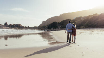 Mature couple walking with arms around on the beach in morning. Loving mature couple having a walk on the sea shore. - JLPSF00469