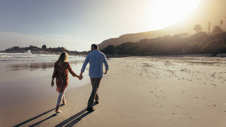 Rear view of romantic senior couple walking hand in hand on the beach. Mature man and woman taking a walk along the sea shore. - JLPSF00470