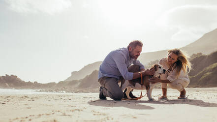 Glückliches reifes Paar streichelt ihren Hund am Strand. Älterer Mann und Frau am Meeresufer mit ihrem Hund am Morgen. - JLPSF00482