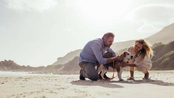 Glückliches reifes Paar streichelt ihren Hund am Strand. Älterer Mann und Frau am Meeresufer mit ihrem Hund am Morgen. - JLPSF00482