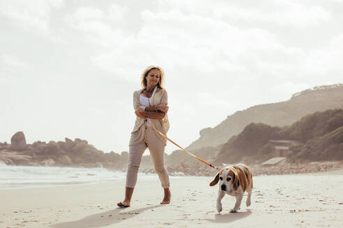 Reife Frau geht mit ihrem Hund am Strand spazieren. Ältere Frau auf Morgenspaziergang mit ihrem Hund. - JLPSF00485
