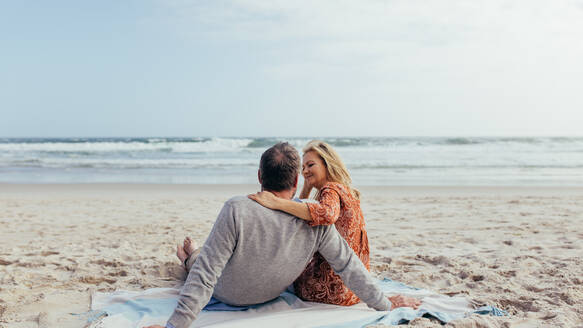 Rückansicht eines reifen Mannes und einer Frau, die zusammen auf einem Strandtuch am Meer sitzen. Romantisches älteres Paar entspannt sich am Strand. - JLPSF00486