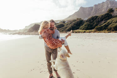 Handsome mature man carrying his wife in his arms on the beach with their pet dog in front. Romantic senior couple with a dog on sea shore. - JLPSF00487