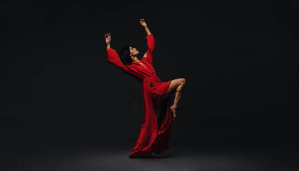 Full length of a young woman dancing in studio. Contemporary female dancer in red dress performing over black background. - JLPSF00495