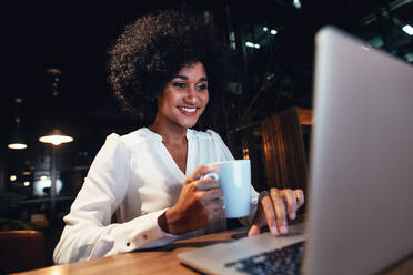 Junge Geschäftsfrau arbeitet am Laptop in der Nachtschicht. Glückliche Frau am Schreibtisch, die eine Tasse Kaffee hält und auf den Laptop schaut. - JLPSF00508