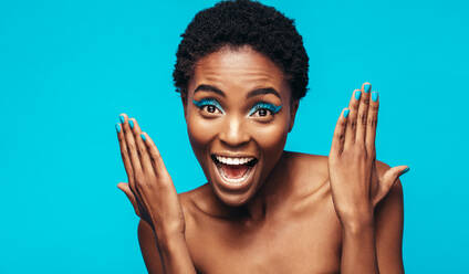 Close up of woman with blue eye shadow and nail paint against blue background. Excited female model showing her vivid makeup. - JLPSF00524