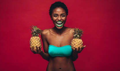 Junge glückliche afroamerikanische Frau, die frische Ananas in der Hand hält und lacht. Modemodell in lebhaftem Make-up, das vor einem roten Hintergrund steht. - JLPSF00544