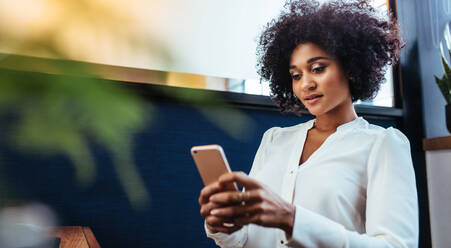 Innenaufnahme einer schönen afrikanischen Geschäftsfrau, die ein Smartphone im Büro benutzt. Eine weibliche Führungskraft bei der Arbeit liest eine Textnachricht auf ihrem Mobiltelefon. - JLPSF00613