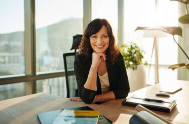 Porträt einer lächelnden Geschäftsfrau, die an ihrem Schreibtisch sitzt. Schöne weibliche Führungskraft in einem modernen Büro, die in die Kamera schaut. - JLPSF00619