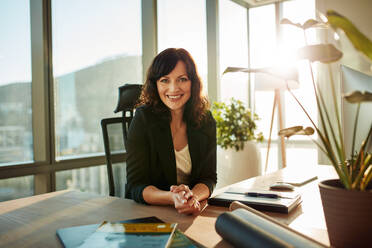 Portrait of female entrepreneur sitting at her desk looking at camera and smiling. Caucasian young woman at her modern workplace. - JLPSF00620