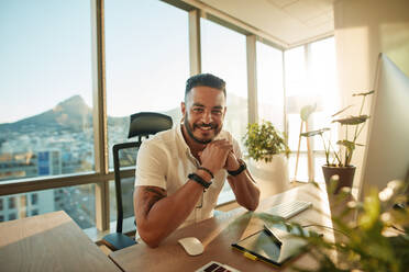 Porträt eines kreativen jungen Mannes im Büro: Ein männlicher Designer sitzt an seinem Schreibtisch und lächelt. - JLPSF00636