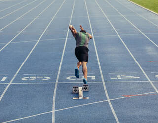 Rückansicht eines Athleten, der seinen Sprint auf einer Allwetter-Laufbahn beginnt. Ein Läufer benutzt einen Startblock, um seinen Lauf auf der Laufbahn zu beginnen. - JLPSF00659