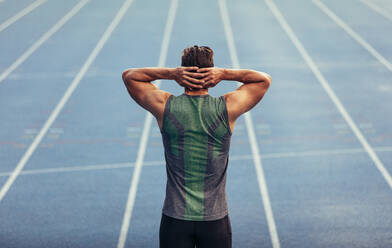 Rear view of an athlete standing on a running track with hands at the back of head. Runner getting ready to race standing on race track. - JLPSF00661