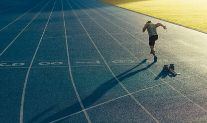 Athlete running on an all-weather running track alone. Runner sprinting on a blue rubberized running track starting off using a starting block. - JLPSF00664