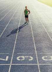Runner standing on running track with hands folded. Athlete wearing earphones with mobile phone fixed in arm band. - JLPSF00681