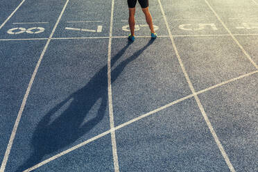 Rear view of legs of athlete standing on an all-weather running track. Shadow of a runner standing at the start line with hands on waist. - JLPSF00683