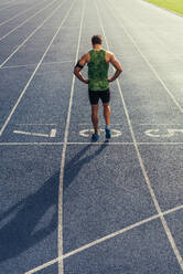 Rear view of an athlete standing on an all-weather running track. Runner standing at the start line with hands on waist. - JLPSF00684