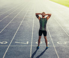 Rear view of an athlete standing on a running track with hands at the back of head. Runner getting ready to run standing at the start line on race track. - JLPSF00686