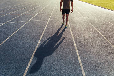 Athlete walking on an all-weather running track listening to music. Shadow of a runner walking on the track. - JLPSF00695