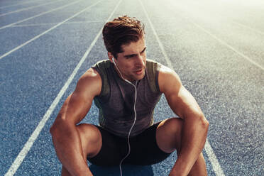 Nahaufnahme eines Sportlers, der auf einer Laufbahn sitzt und Musik hört. Ein Läufer mit Kopfhörern sitzt und entspannt auf der Laufbahn. - JLPSF00698