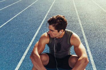 Close up of an athlete sitting on a running track listening to music. Runner wearing earphones sitting and relaxing on the running track looking away. - JLPSF00703