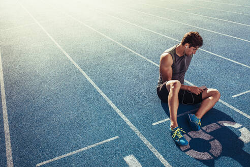 Athlete sitting on a running track listening to music. Runner wearing earphones and using mobile phone sitting at the start line on the running track. - JLPSF00706