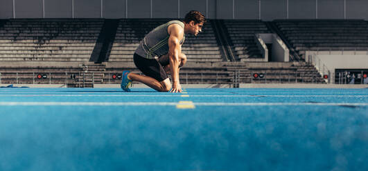 Side view of an athlete on his marks to start off the run. Runner practicing his sprint on an all-weather running track in a stadium. - JLPSF00709