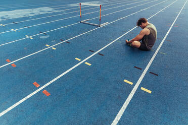 Athlete doing stretching exercises sitting on the running track. Runner sitting beside a hurdle on running track with feet joined together. - JLPSF00716