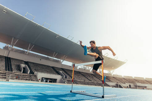 Läufer, der bei einem Leichtathletikwettkampf über eine Hürde springt. Athlet, der einen Hürdenlauf in einem Stadion absolviert. - JLPSF00717