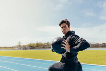 Runner doing a medicine ball workout on running track. Athlete using a medicine ball for fitness training at a track and field stadium. - JLPSF00722