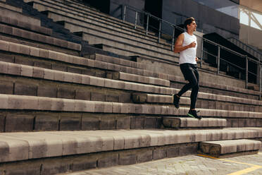 Läufer, der im Rahmen seines körperlichen Trainings die Stufen der Stadiontribüne hinunterläuft. Junger Mann, der die Treppe zum Stadion hinunterläuft. - JLPSF00729