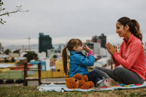 Seitenansicht einer fröhlich lächelnden Frau und eines Mädchens, die in einem Park Kuchen spielen. Ein kleines Mädchen spielt mit ihrem Kindermädchen ein Klatschspiel im Freien. - JLPSF00736