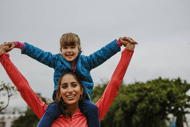 Woman carrying a girl on her shoulder outdoors. Small girl sitting on shoulder of her nanny and smiling. - JLPSF00738