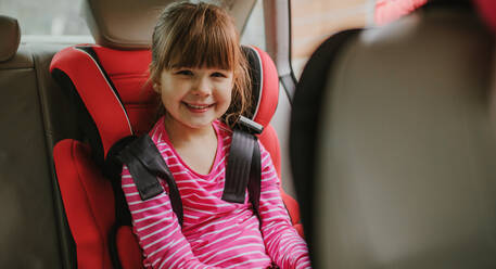 Lächelndes Mädchen, das mit einem Sicherheitsgurt auf dem Rücksitz eines Autos angeschnallt ist. Niedliches Mädchen, das in ihrem Autositz angeschnallt ist. - JLPSF00739
