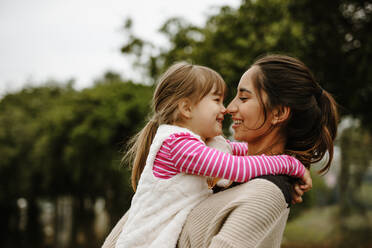 Seitenansicht einer Frau, die ein kleines Mädchen trägt und im Freien lächelt. Babysitterin mit einem Mädchen im Park. - JLPSF00741