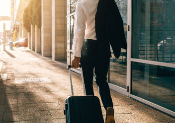 Rear view of young businessman walking outside public transport building with suitcase. Business traveler pulling suitcase on city street. - JLPSF00747