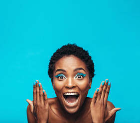 Excited young woman with vibrant eye shadow and nail paint. Female model showing her vivid makeup against blue background. - JLPSF00759