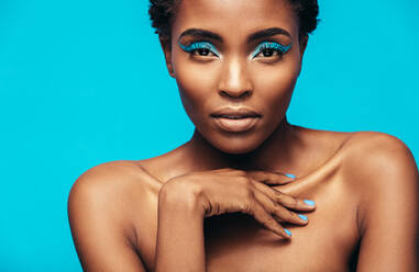 Nahaufnahme einer sinnlichen afrikanischen Frau mit lebhaftem Make-up vor blauem Hintergrund. Weibliches Modell mit schöner Haut schaut in die Kamera. - JLPSF00760