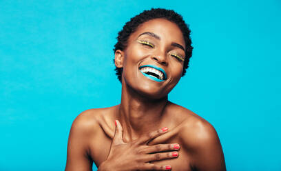 Nahaufnahme einer jungen Frau mit buntem Make-up, die vor einem blauen Hintergrund lächelt. Schönheitsaufnahme eines afrikanischen weiblichen Modells mit lebhaftem Make-up. - JLPSF00766
