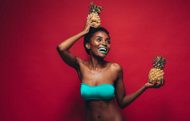 Schöne junge Frau mit Ananas in der Hand, die wegschaut. Lachendes weibliches Model mit Ananas, das über einen roten Hintergrund lacht. - JLPSF00770