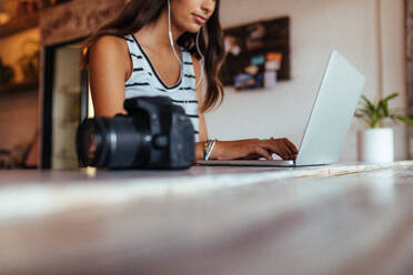 Frau Bloggerin mit Laptop zu Hause mit Kopfhörern. Frau sitzt mit DSLR-Kamera auf dem Tisch arbeiten auf ihrem Laptop-Computer. - JLPSF00776