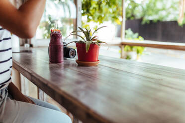 Eine Frau sitzt am Fenster mit einem Smoothie auf dem Tisch. Ein Smoothie-Glas steht auf einem Tisch neben einer Kamera und einem Blumentopf. - JLPSF00794