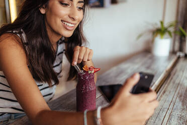Woman taking a selfie with a smoothie using a mobile phone for her food blog. Food blogger shooting photos for her blog at home. - JLPSF00797