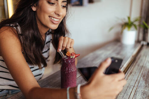 Frau, die mit einem Mobiltelefon ein Selfie mit einem Smoothie für ihren Food-Blog macht. Food-Bloggerin, die zu Hause Fotos für ihren Blog macht. - JLPSF00797