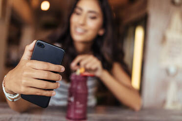 Woman taking a selfie with a smoothie placed on the table using a mobile phone for her food blog. Food blogger shooting photos for her blog at home. - JLPSF00798