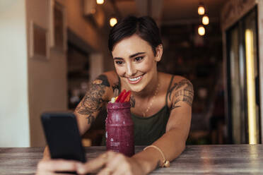 Lächelnde Frau, die mit einem Mobiltelefon ein Selfie mit einem Smoothie vor sich aufnimmt, den sie für ihren Food-Blog verwendet. Food-Bloggerin, die zu Hause Fotos für ihren Blog schießt. - JLPSF00801