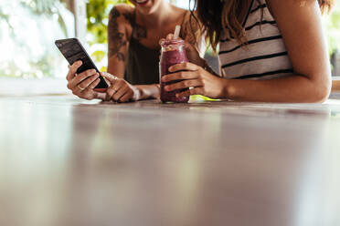 Nahaufnahme von zwei Frauen, die in einem Restaurant sitzen und auf ihr Handy schauen. Eine Frau zeigt ihr Handy, während eine andere Frau ein Smoothie-Glas hält. - JLPSF00820