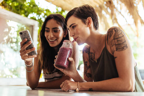 Nahaufnahme von zwei Frauen, die in einem Restaurant sitzen und auf ihr Handy schauen. Eine Frau zeigt ihr Handy, während die andere Frau einen Smoothie genießt. - JLPSF00821