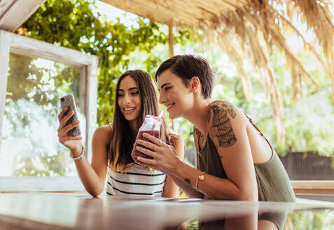 Zwei Frauen sitzen in einem Restaurant und schauen auf ihr Handy. Eine Frau zeigt ihr Handy, während die andere Frau einen Smoothie genießt. - JLPSF00822