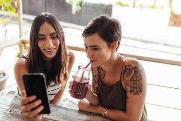 Zwei Frauen sitzen in einem Restaurant und schauen auf ein Handy. Eine Frau zeigt ihr Handy, während die andere Frau einen Smoothie genießt. - JLPSF00823